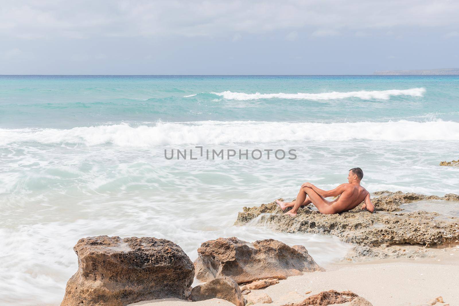 Male naturist lying on wet rocky shore near waving sea during vacation on Formentera island in Spain