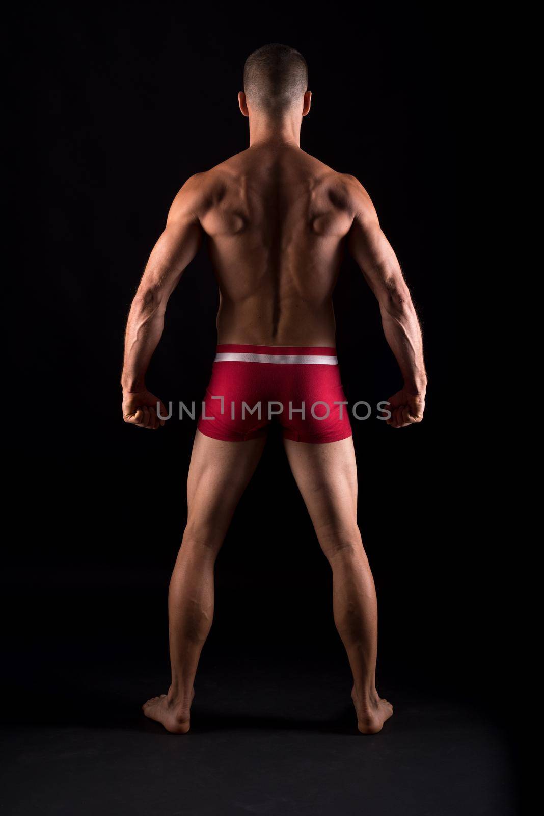 Vertical studio portrait of the back of a strong adult male in pants standing with black background