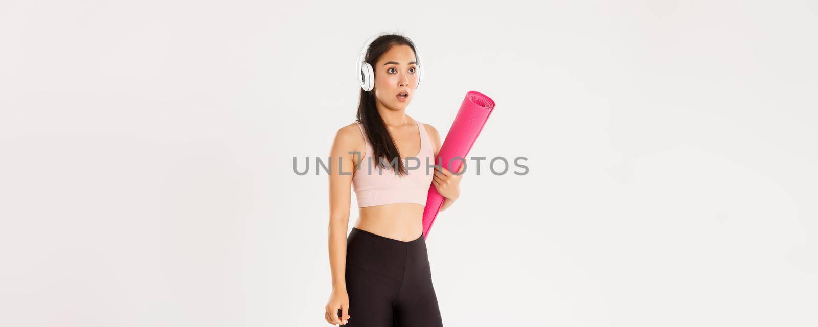 Sport, wellbeing and active lifestyle concept. Shocked and startled asian girl in headphones, holding rubber mat, stop and drop jaw as looking at logo or closed sign, covid-19 quarantine measures by Benzoix