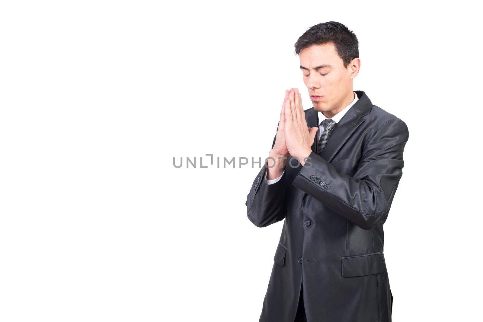 Thoughtful adult male with closed eyes wearing black formal suit praying with clasped hands against white background