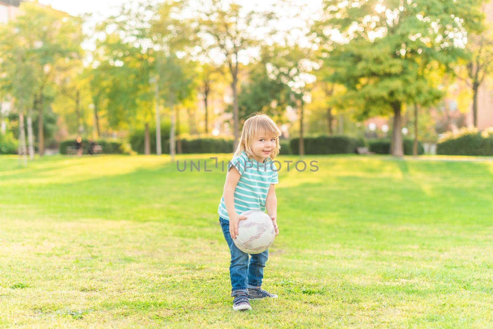 Full length view of a little boy standing on grass in park holding a ball in a sunny day.