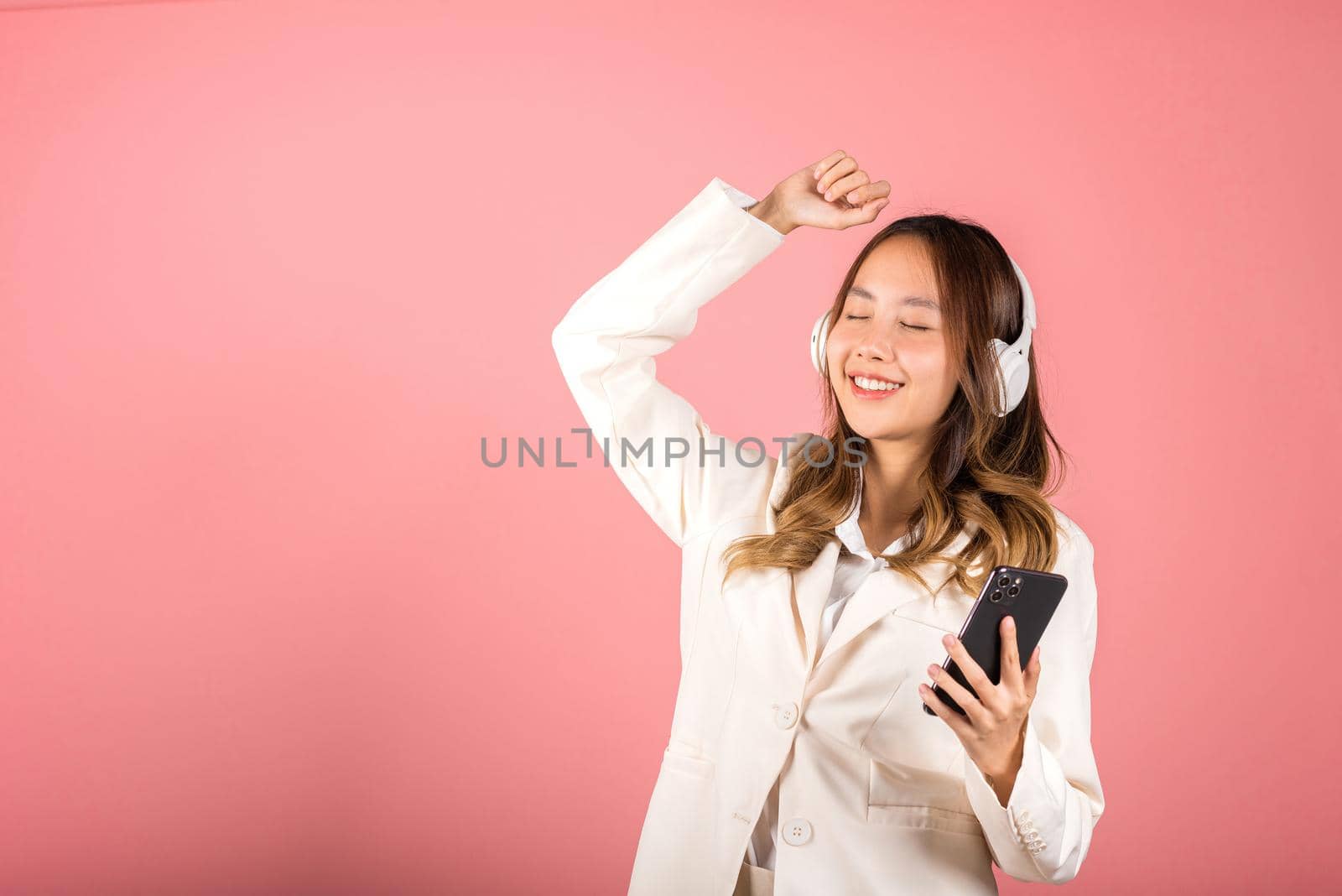 Woman excited smiling listening to music radio in bluetooth headphones and raise hand dancing on smartphone studio shot isolated white background, Happy Asian young female online app in mobile phone