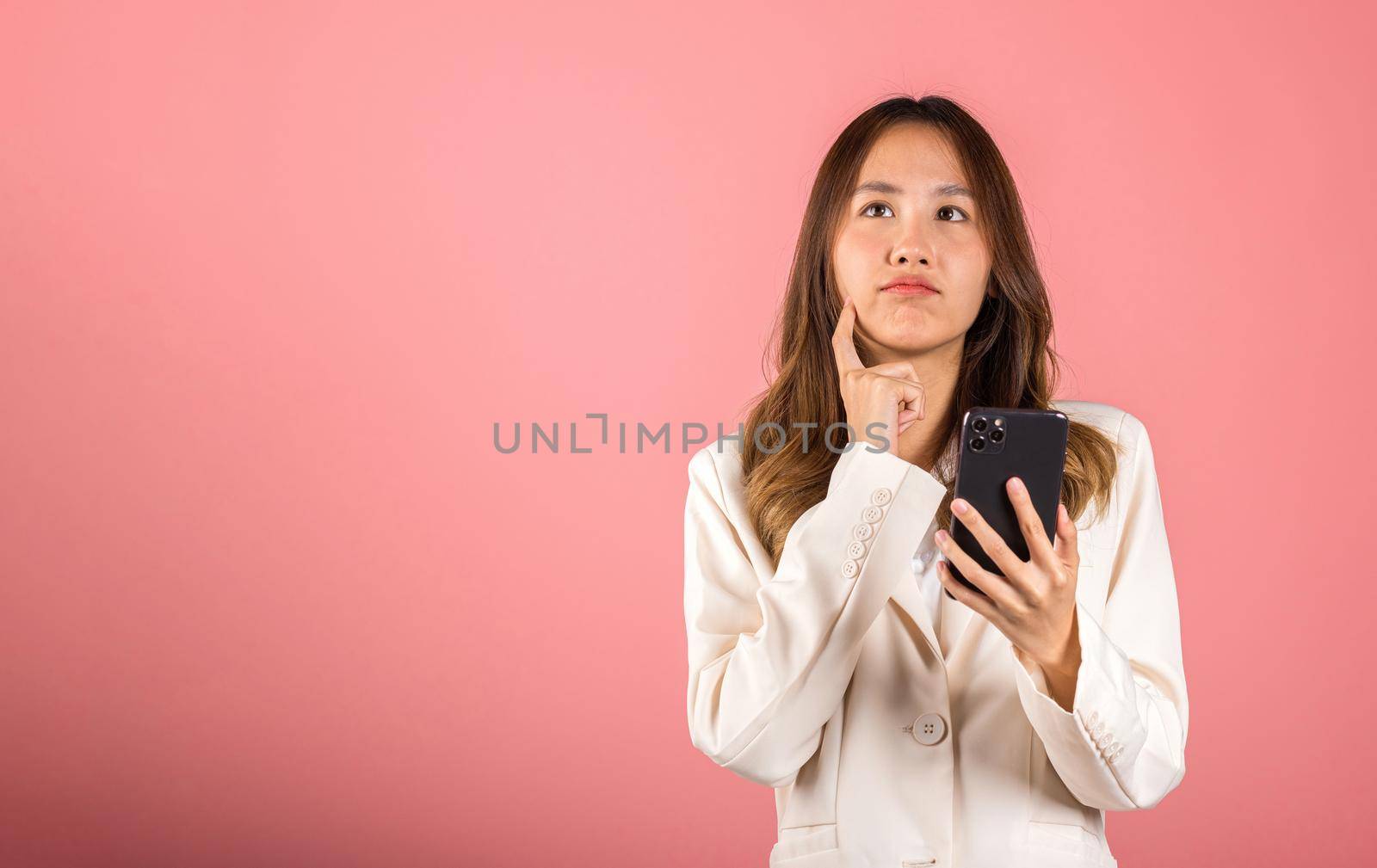 Happy Asian portrait beautiful cute young woman excited smiling holding mobile phone and think idea finger touch face, studio shot isolated on pink background, female using smartphone making gesture