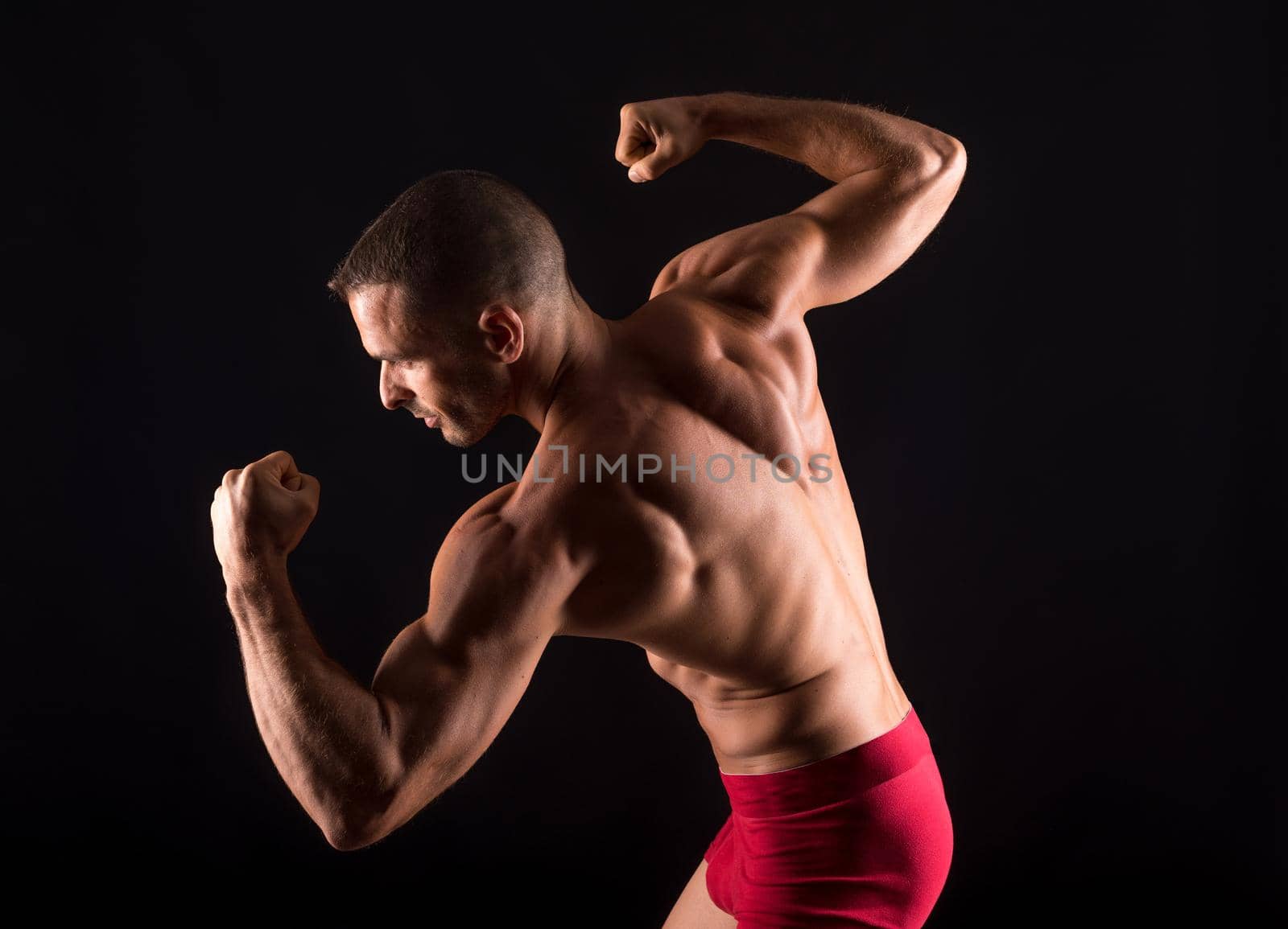 Muscular man on his back and shirtless, sticking out biceps. Mid shot. Black background.