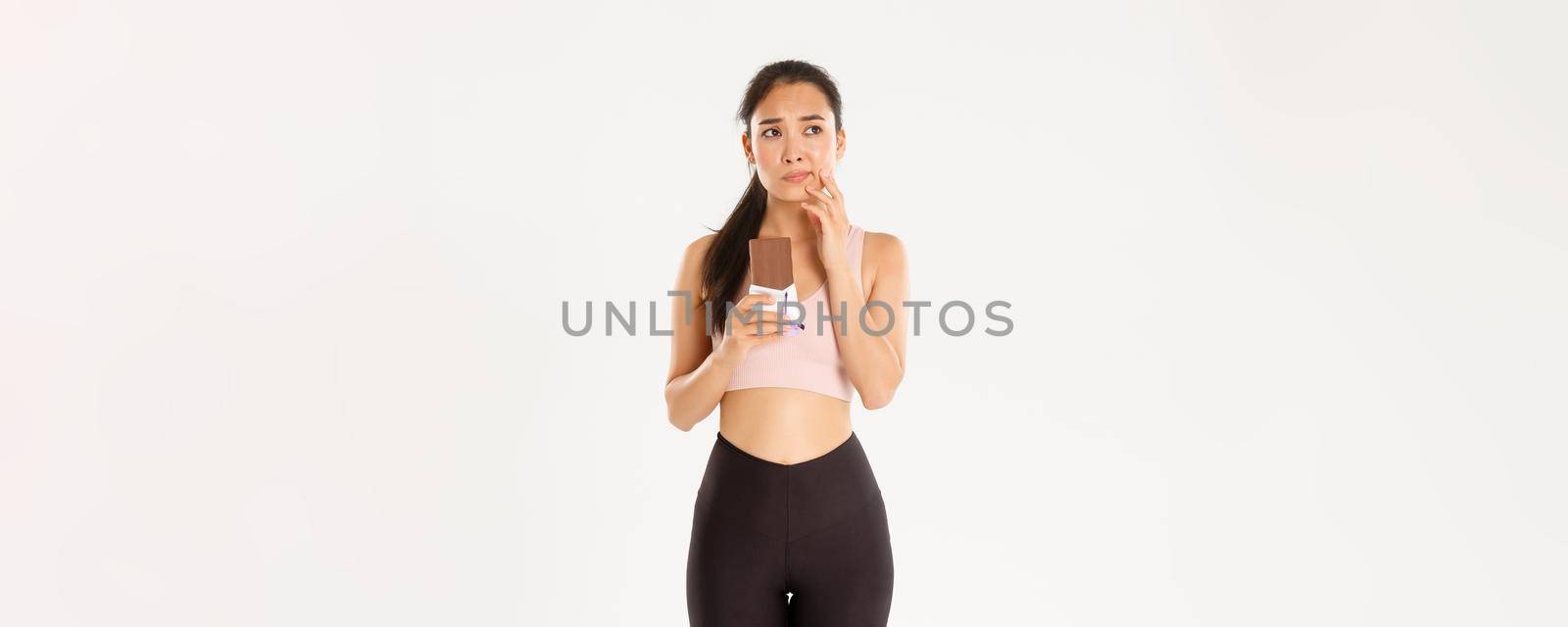 Sport, wellbeing and active lifestyle concept. Doubtful and indecisive asian fitness girl holding chocolate bar and thinking about eating it, looking away hesitant, worried about calories.