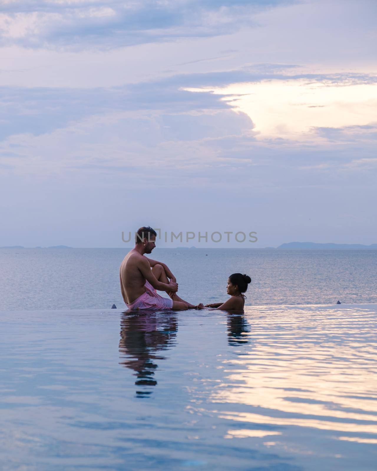 couple men and women relaxing on the edge of a pool during sunset by ocean at a luxury vacation. Men and women watching the sunset in the swimming pool