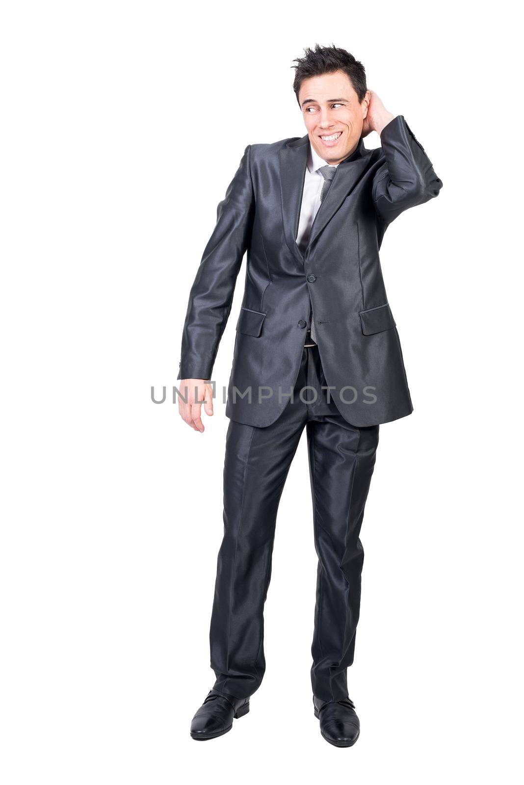 Full body of young elegant shy guy with dark hair in classy suit smiling and looking away while touching head standing in white studio