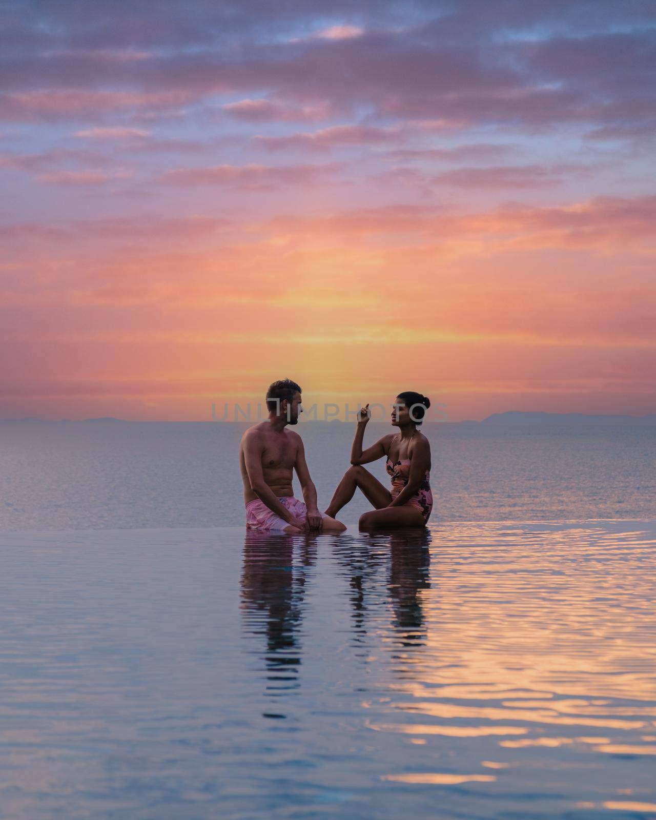 couple men and women relaxing on the edge of a pool during sunset by ocean at a luxury vacation. Men and women watching the sunset in the swimming pool