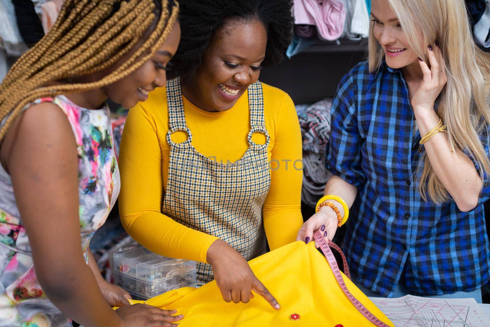 Creative dark-skinned girls and one European woman match buttons to a selected sewing fabric. Two African girls and a European woman in a sewing studio.