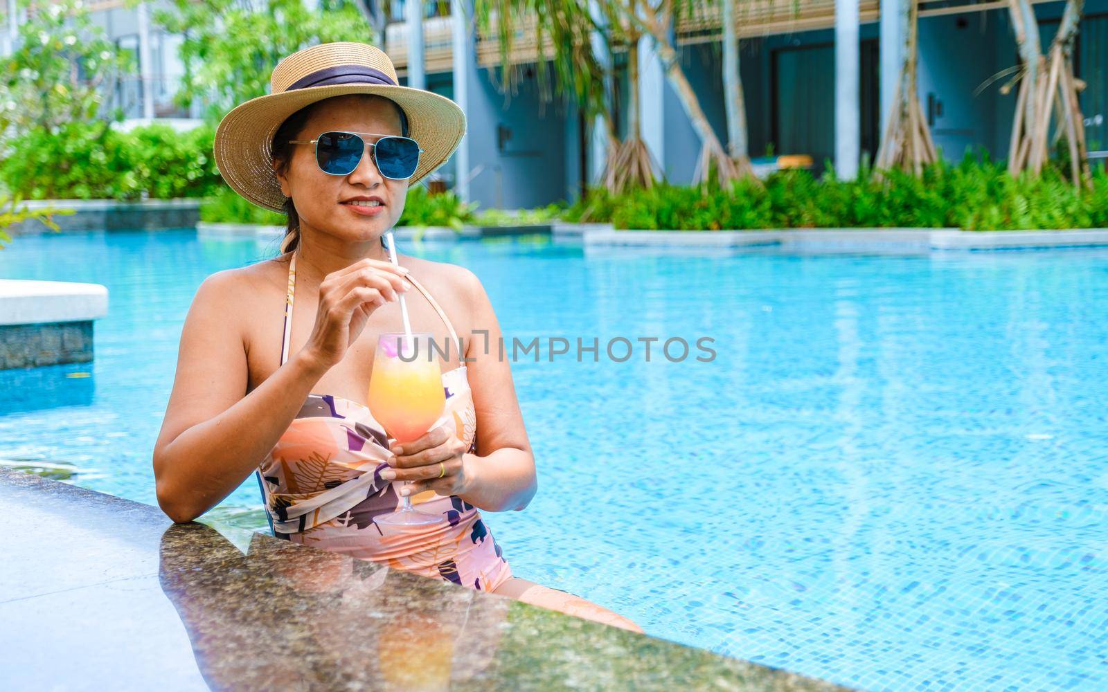 Asian women with a hat relaxing in the swimming pool with a cocktail in hand, women swimming pool banner holiday vacation concept, Asian women in blue swimming pool luxury vacation