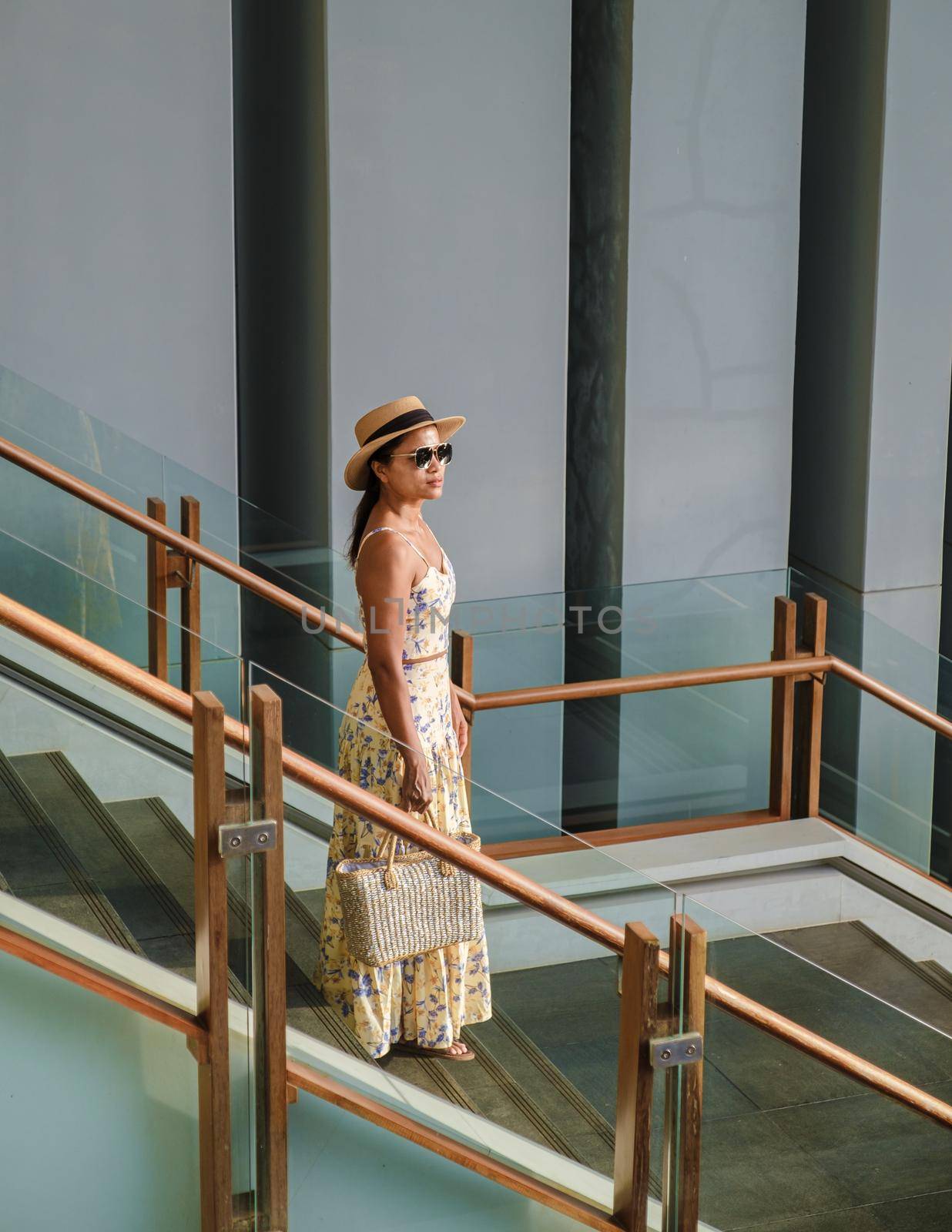 Asian women with hat walking down from stairs with on the back a grey building during sunset by fokkebok