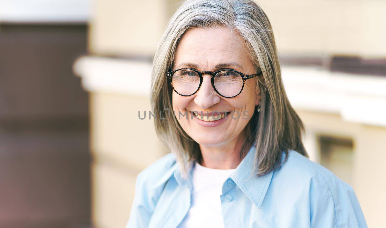 Charming grey hair mature woman wearing eye glasses standing outdoors enjoying vacation traveling around world at retirement. Mature woman with perfect skin wearing blue shirt.