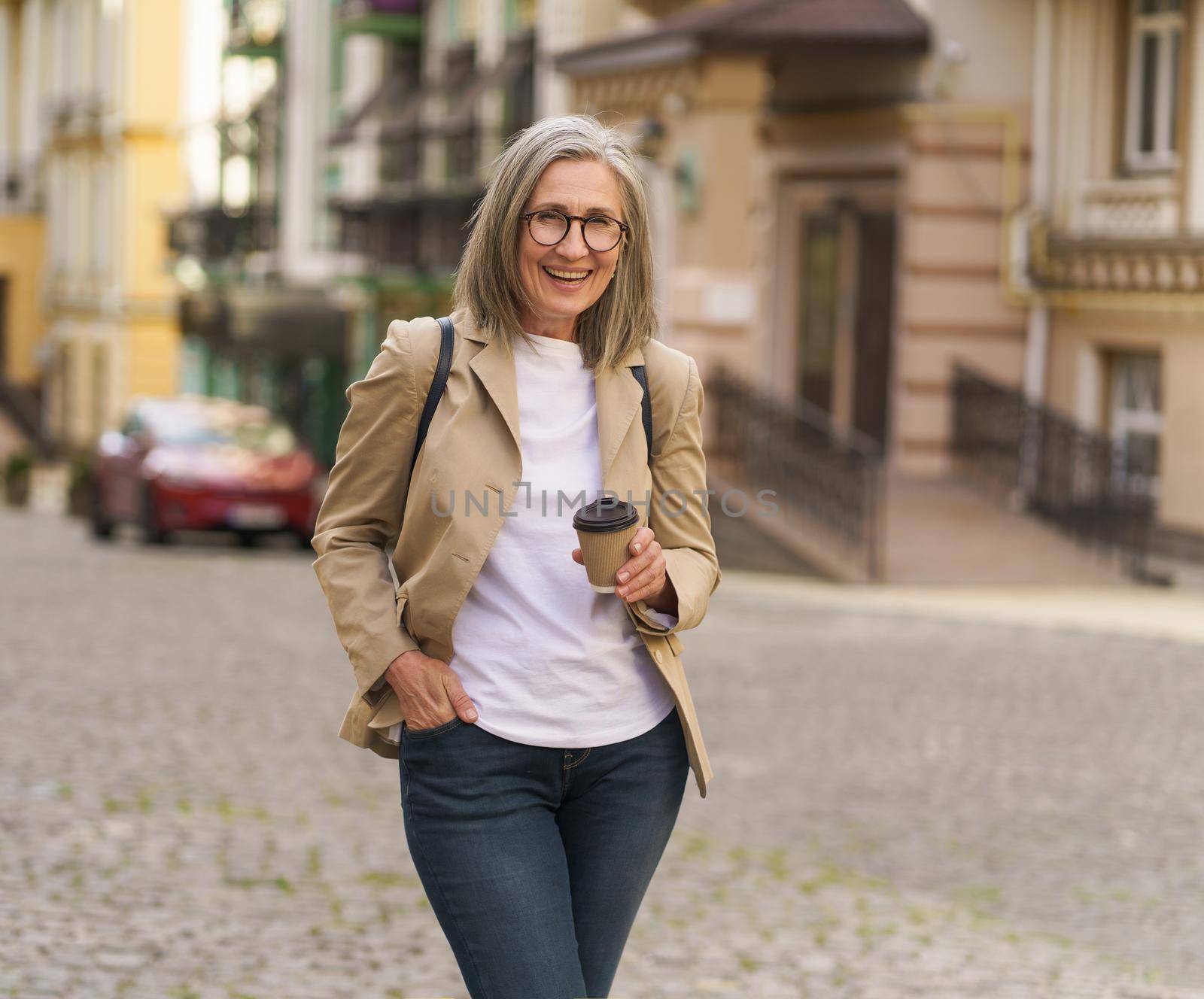 Excited mature businesswoman in casual jacket and denim jeans hold paper cup with hot coffee standing outdoors of old town. Senior woman spend traveling in European city enjoying cup of tea, coffee.