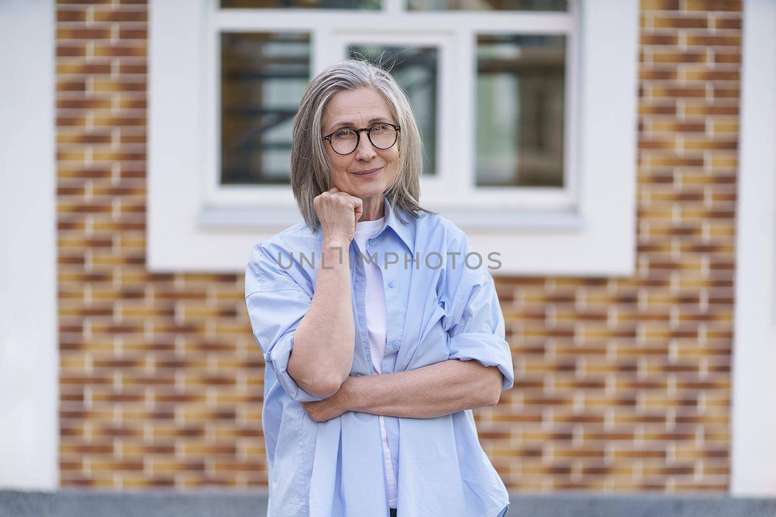 Mature grey hair woman wearing glasses standing outdoor traveling around world at retirement. Senior woman meeting her family. Mature woman with perfect skin wearing blue shirt standing outside.