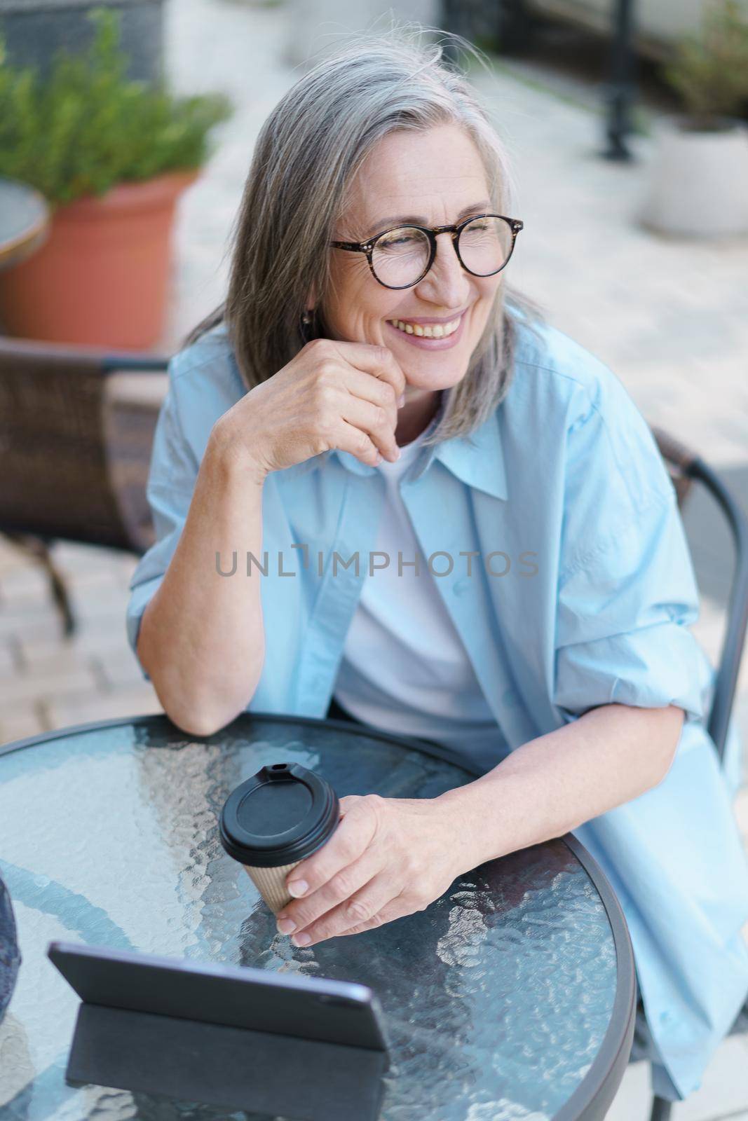 Grey haired mature European smiling woman drink coffee using digital tablet sitting at table of street cafe. Mature silver hair woman working outdoors city cafe, wearing white t-shirt and blue shirt.