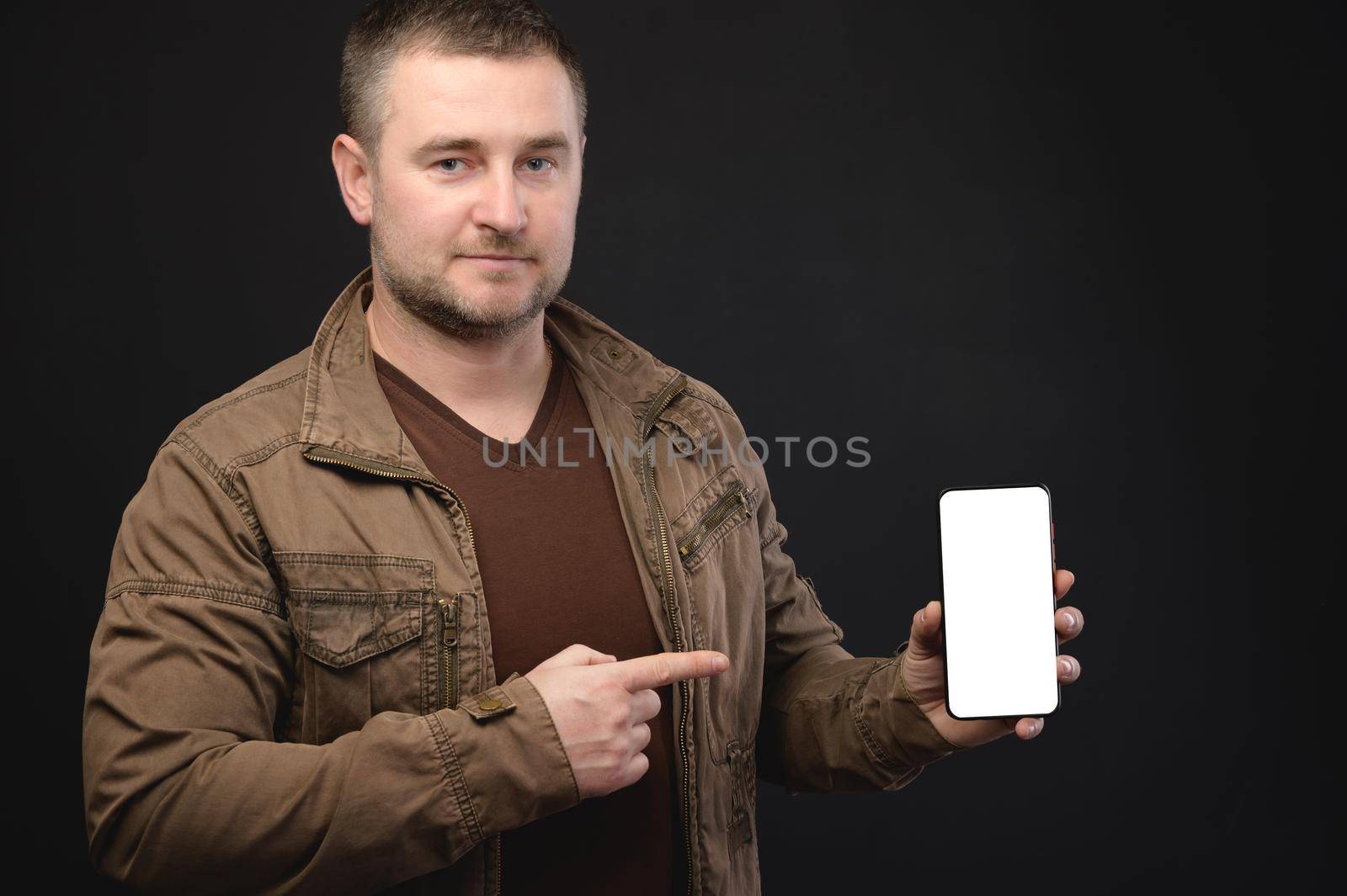 Mature middle-aged man in casual clothes presenting an application on a smartphone while holding in his hand showing a cut-out mockup screen. studio portrait.