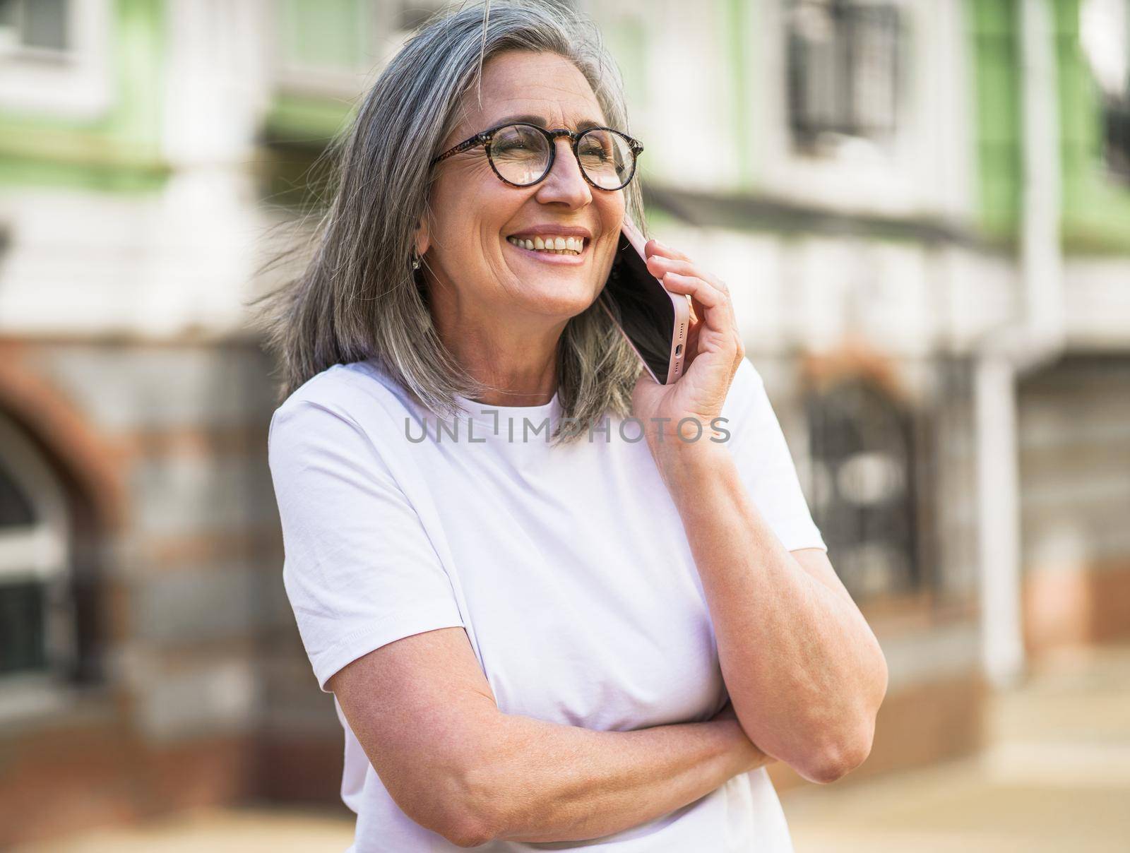 Mature grey hair business woman in eye glasses talking on the phone standing outdoors of the streets of old urban city wearing white t-shirt. Silver hair woman outdoors by LipikStockMedia