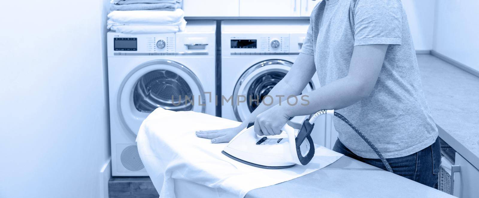 Young Woman ironing white shirt on board in laundry room with washing machine on background by Mariakray