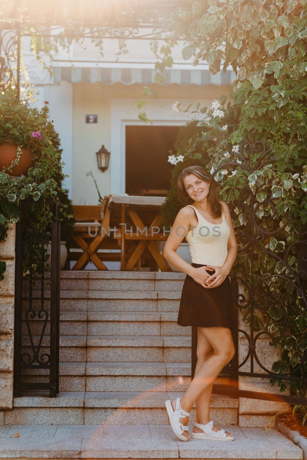 Girl Tourist Walking Through Ancient Narrow Street On A Beautiful Summer Day In MEDITERRANEAN MEDIEVAL CITY, OLD TOWN BUDVA, MONTENEGRO. Young Beautiful Cheerful Woman Walking On Old Street At Tropical Town. Pretty Girl Looking At You And Smiling by Andrii_Ko