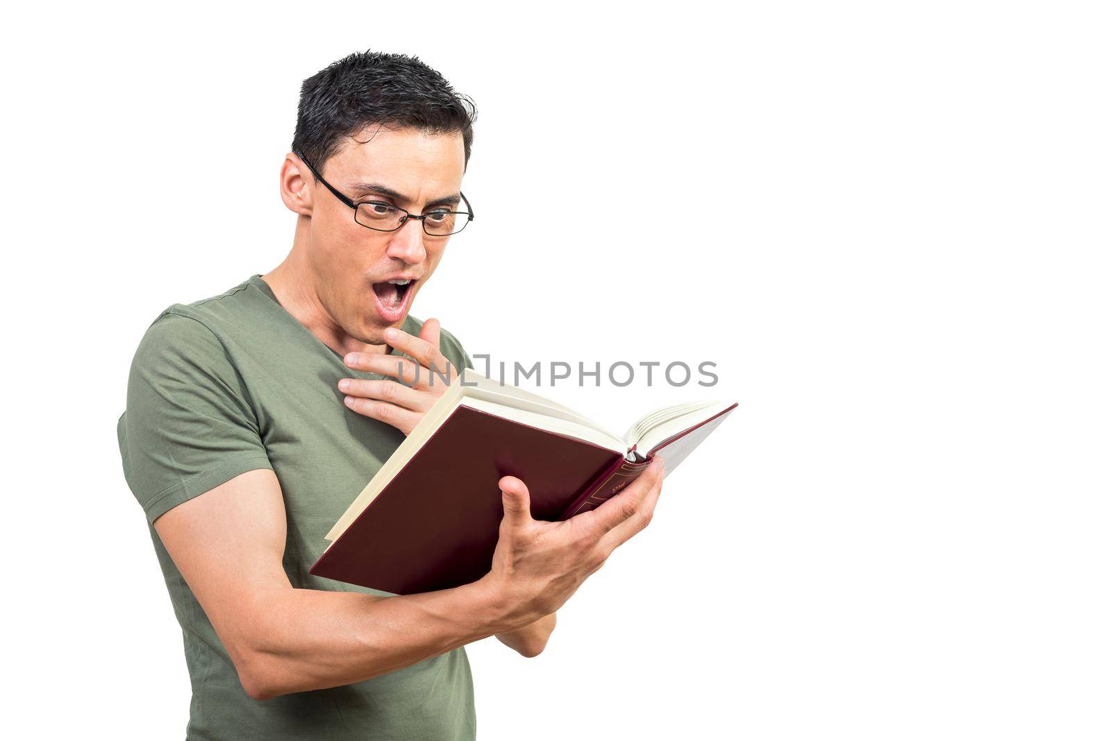 Clever male in green t shirt and glasses touching chin and reading fascinating book with opened mouth against white background