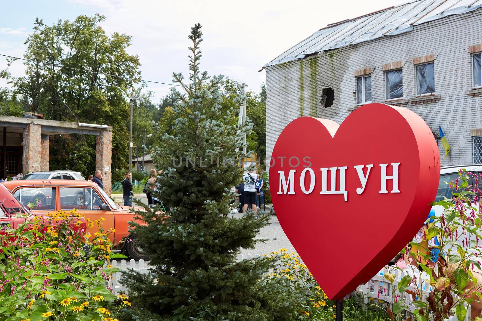 Moshchun, Ukraine - August 25, 2022: People take humanitarian aid from volunteers. The trace from a tank shot on the house. Words on the sign: Moshchun by sarymsakov