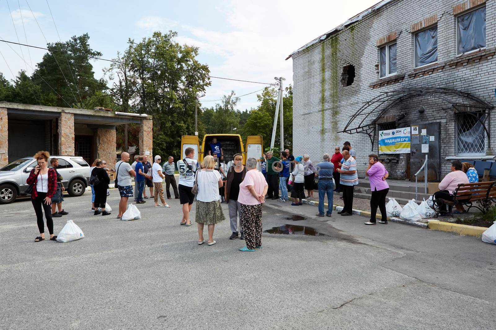 Moshchun, Ukraine - August 25, 2022: People take humanitarian aid from volunteers. The trace from a tank shot on the house. Words on the sign: Moshchun.