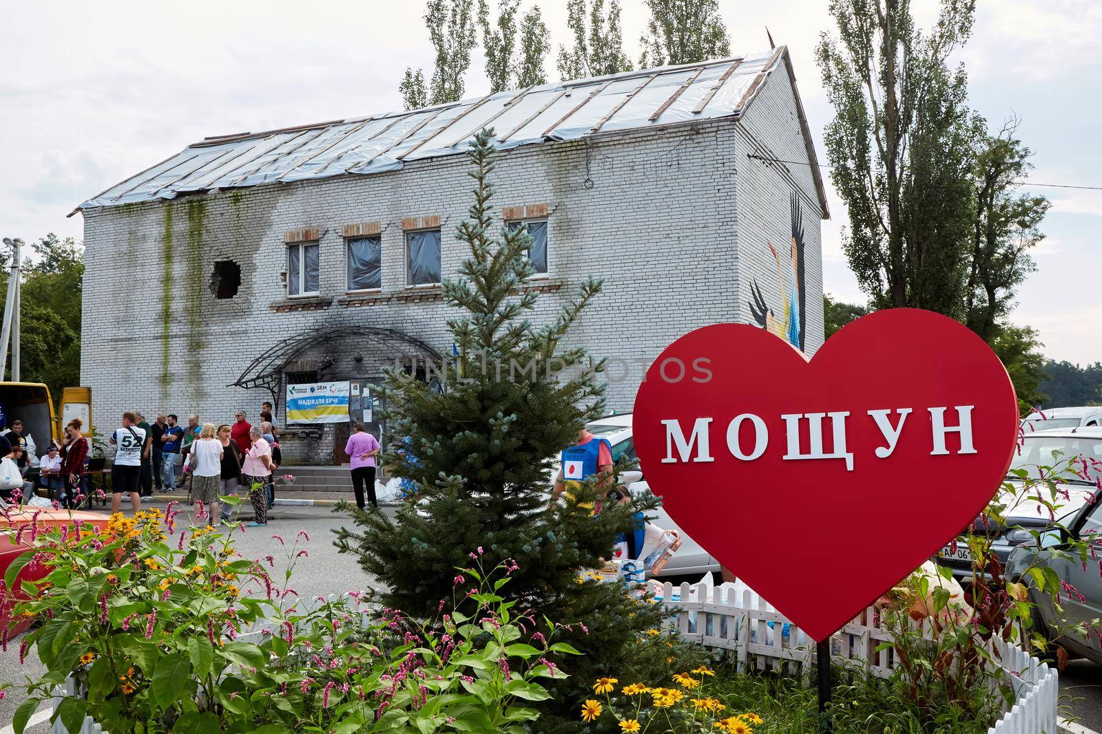Moshchun, Ukraine - August 25, 2022: People take humanitarian aid from volunteers. The trace from a tank shot on the house. Words on the sign: Moshchun by sarymsakov