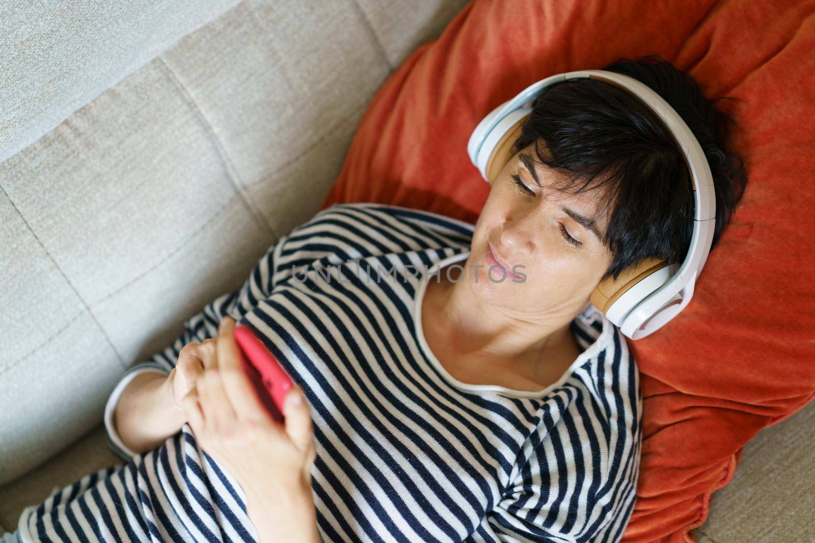 Middle-aged woman consulting her smartphone while listening to music with headphones lying on the couch.