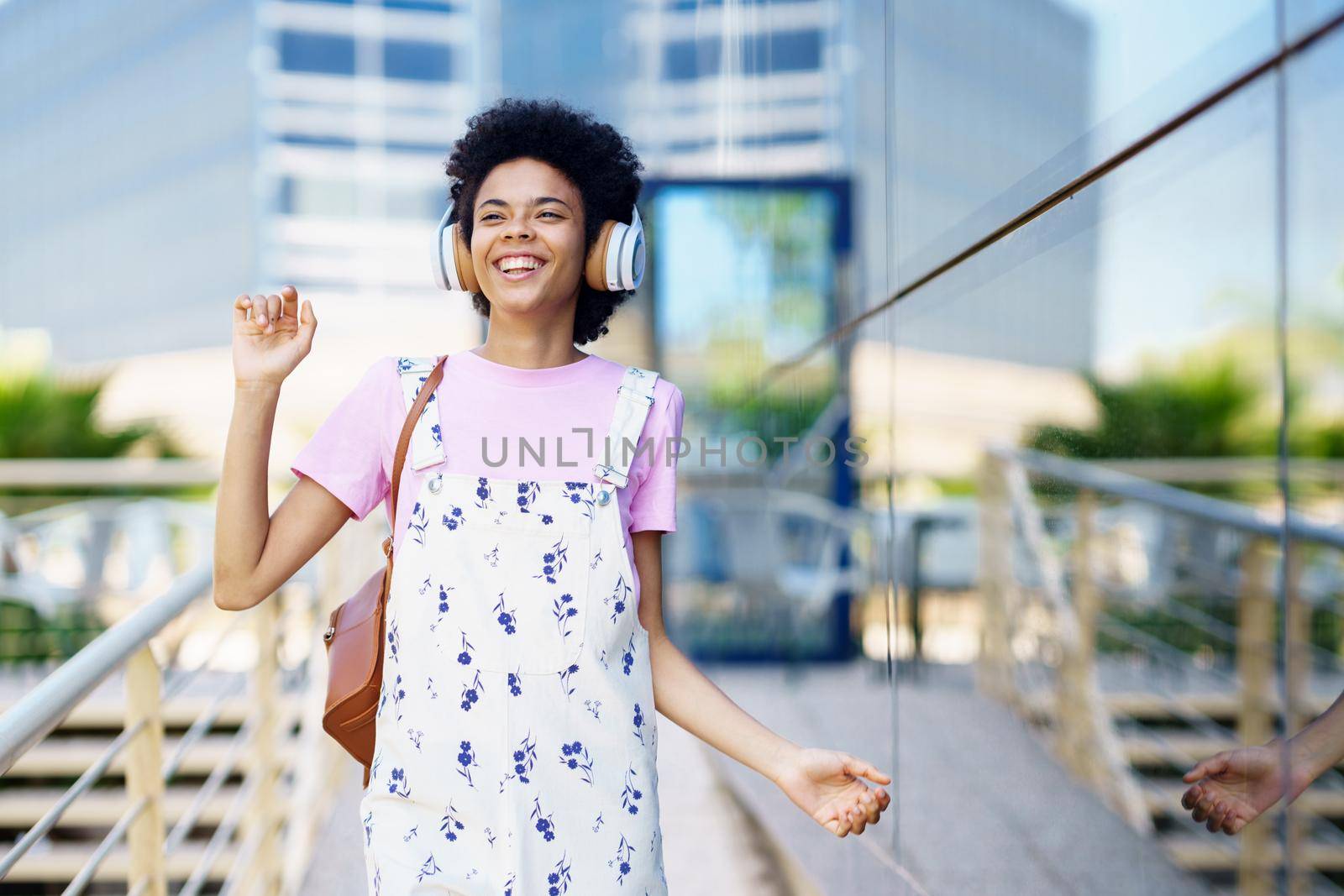 Smiling African American female listening to music in wireless headphones while standing on street near building with glass wall in city