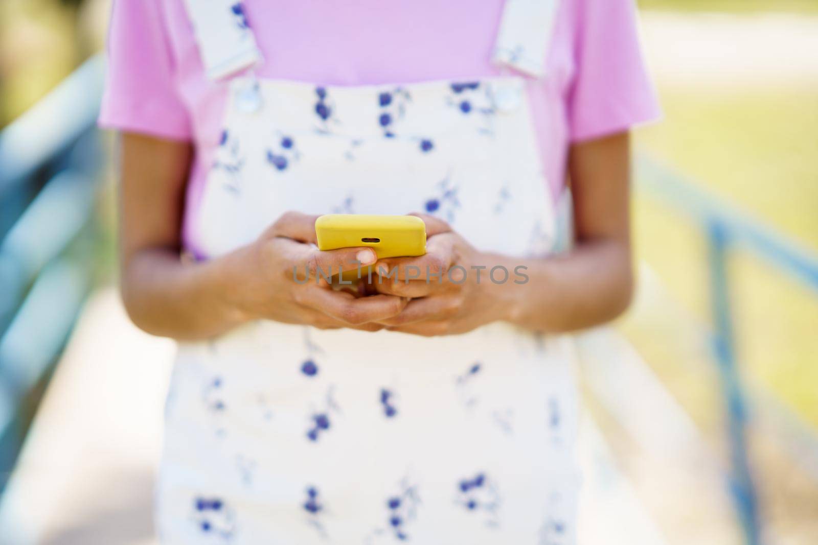 Crop unrecognizable female in overall texting message on cellphone while standing on pathway on street against blurred background on summer day