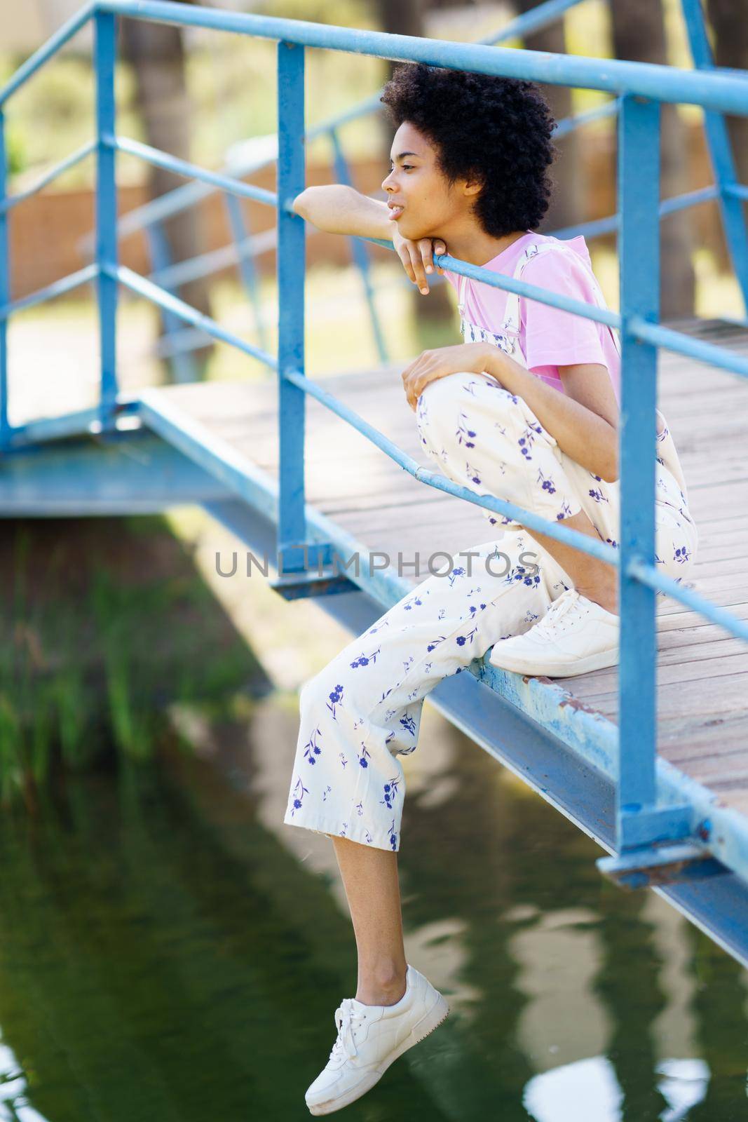 Full body side view of focused African American female sitting on wooden footbridge over lake on summer day in park