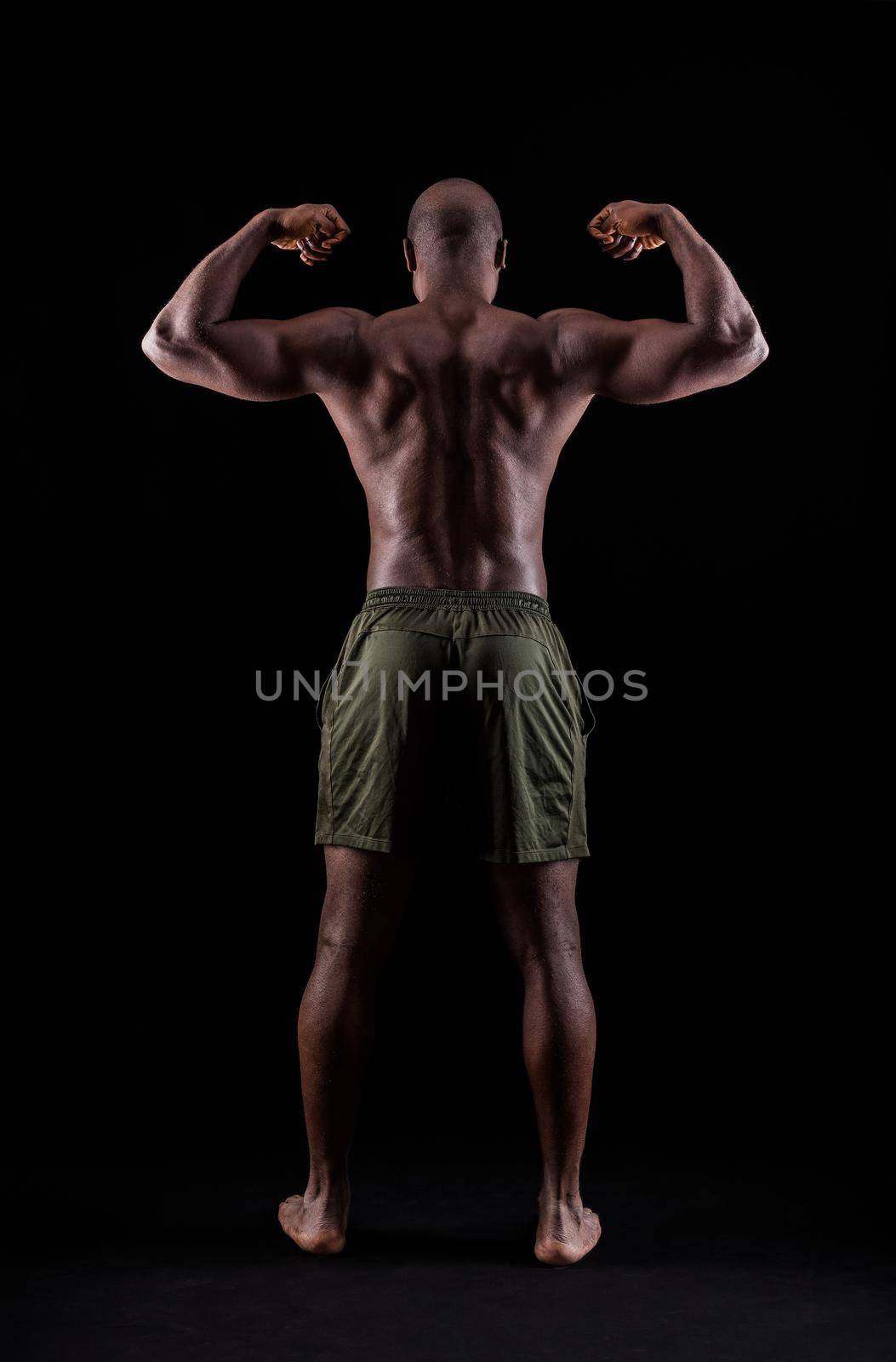 Rear view of an African American muscular man flexing arm muscles on a black background. Athlete adult man standing with shorts in a studio.