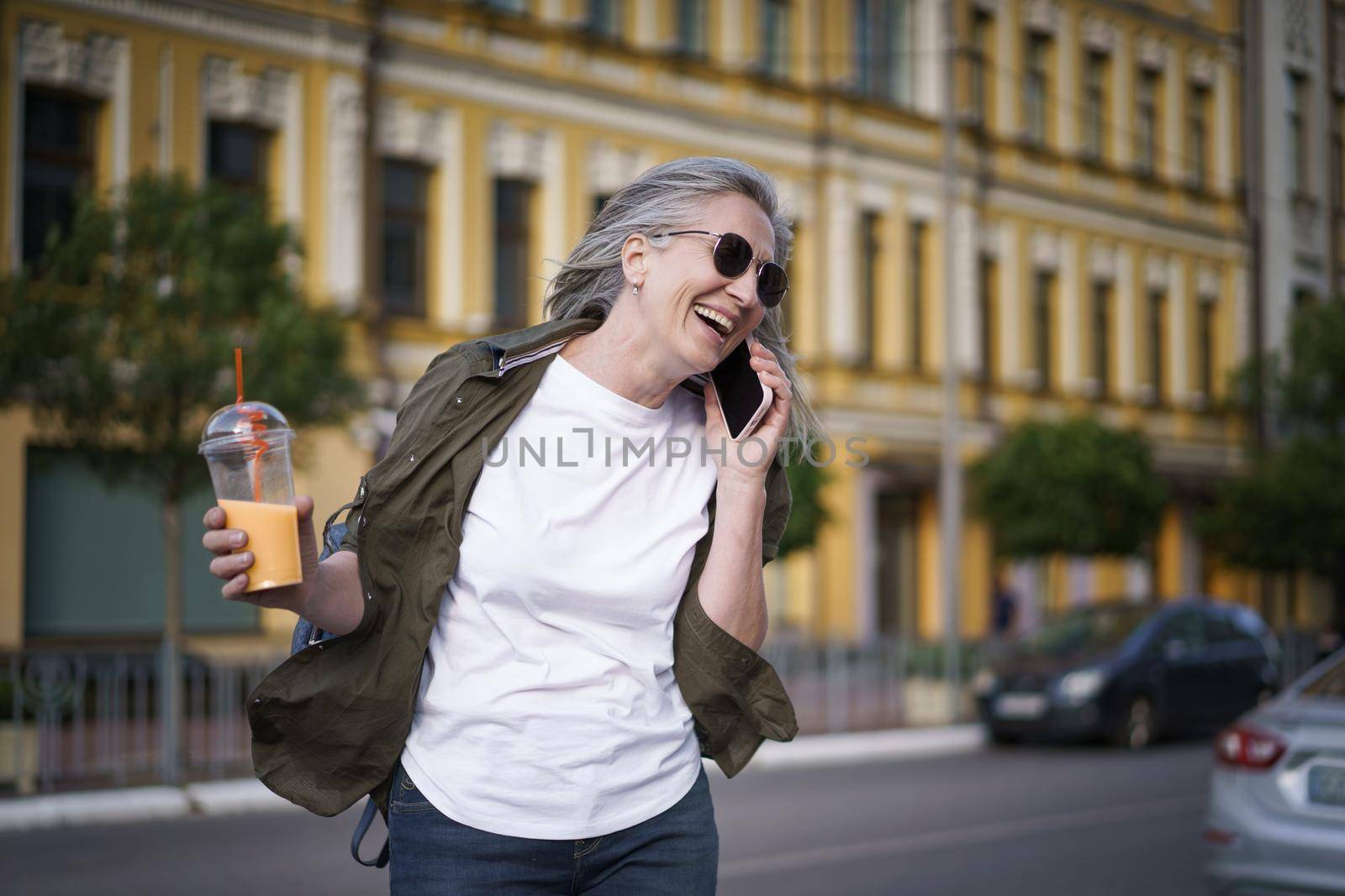 Happy mature European woman laughing talking on the phone enjoying free time after work or traveling having juice on the go using plastic cup in city background. Enjoying life mature woman.