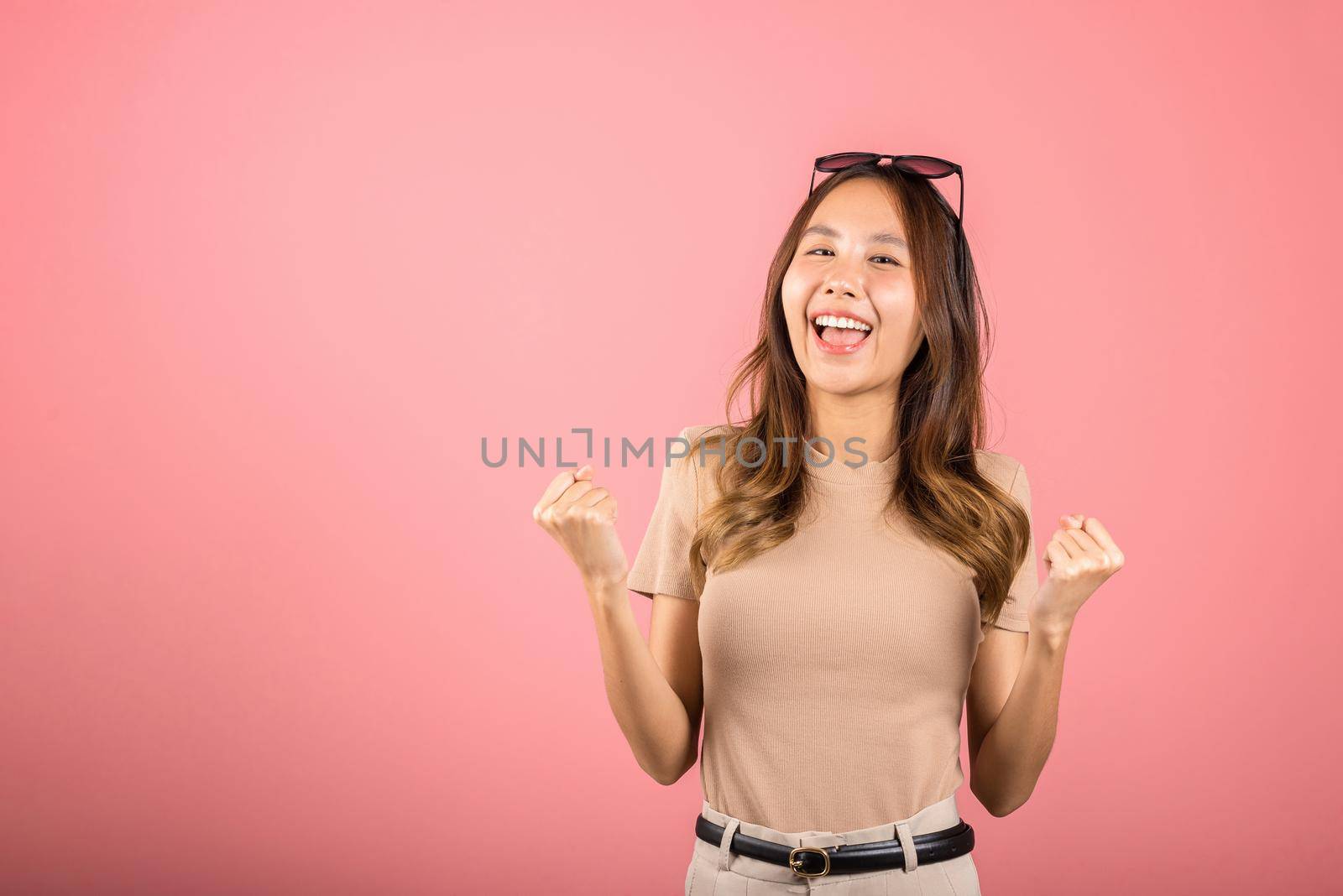 Happy Asian portrait beautiful cute young woman wear glasses makes raised hand up celebrating her winning success gesture, studio shot isolated pink background, Female excited say yes! with copy space