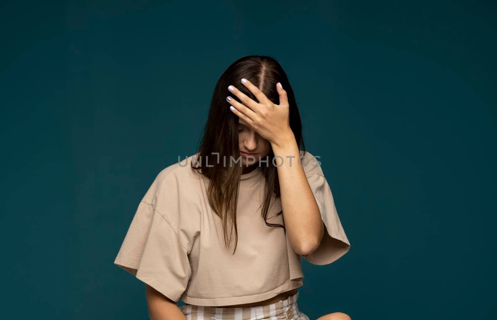 Portrait of stressed beautiful brunette woman in a beige t-shirt holding her head with hands having headache over cyan wall. Attractive female suffering from head pain