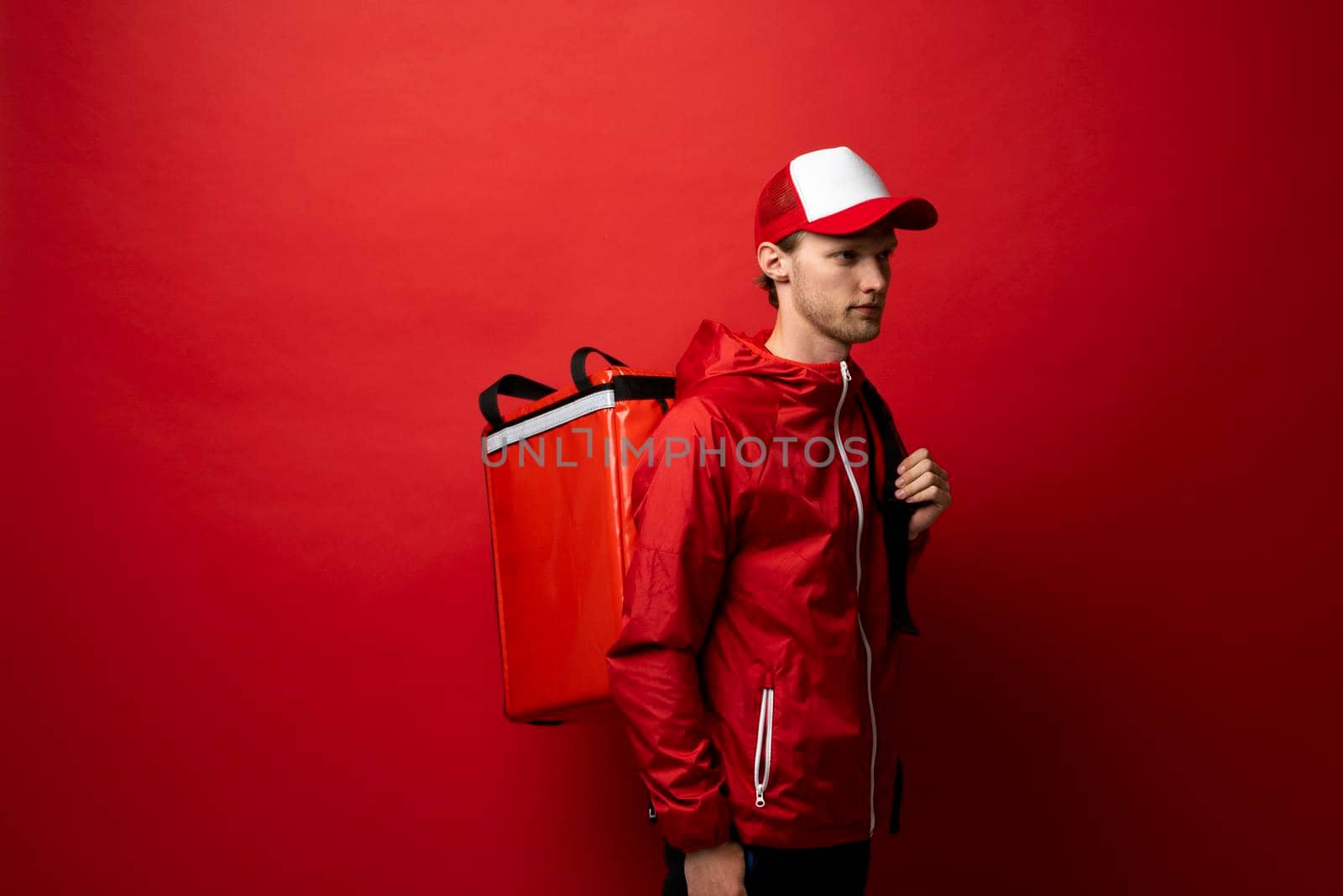 Delivery man in a red jacket and cap carry red thermal backpack full of food, groceries to the clients. Food delivery service