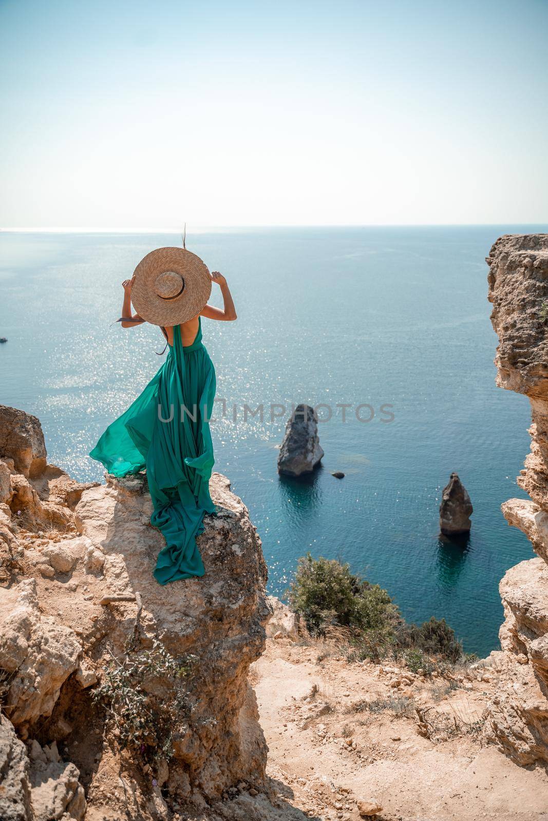 A girl with loose hair in a long mint dress descends the stairs between the yellow rocks overlooking the sea. A rock can be seen in the sea. Sunny path on the sea from the rising sun.