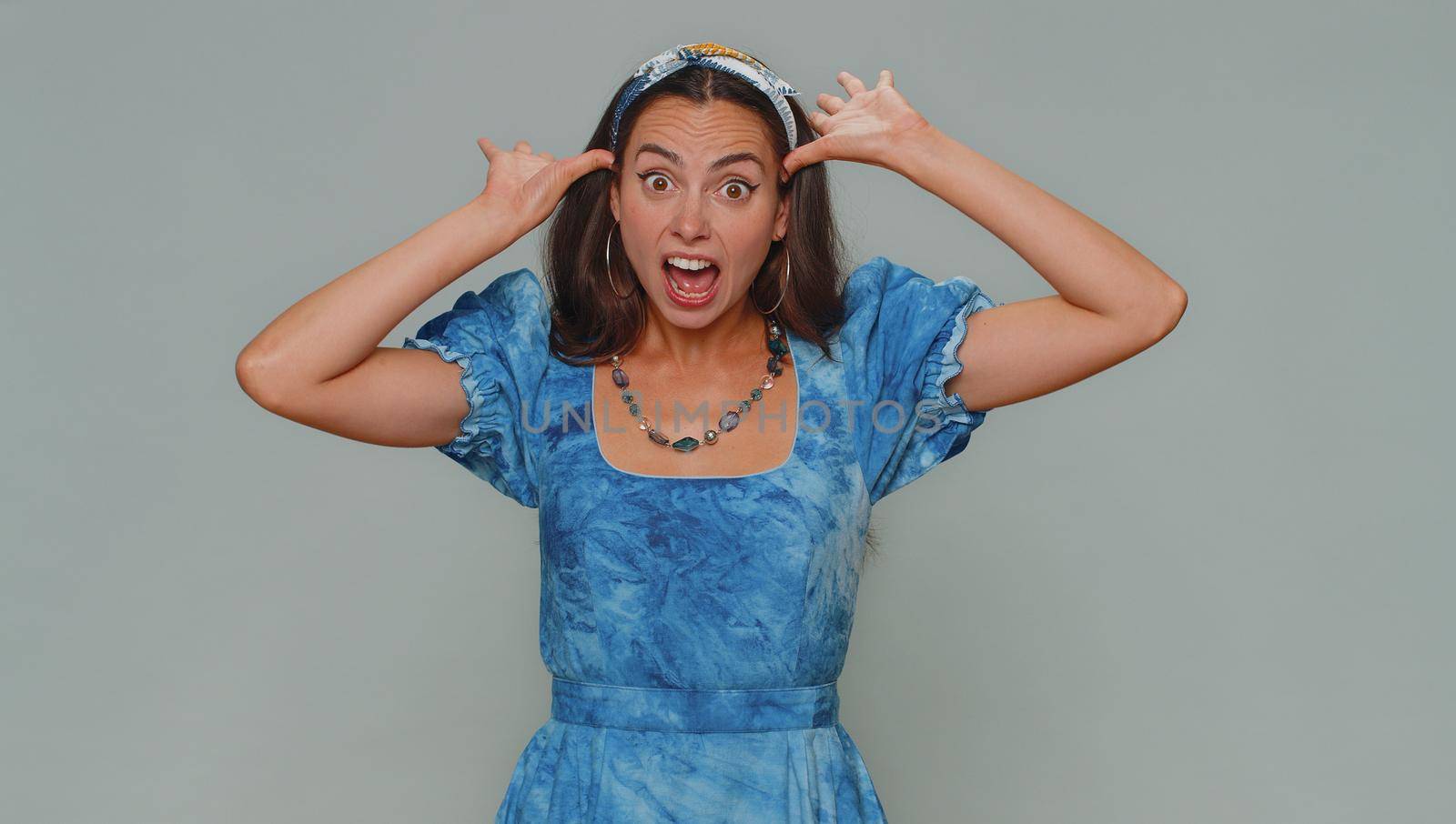 Lovely pretty funny woman in dress making playful silly facial expressions and grimacing, fooling around, showing tongue. Adult stylish female girl isolated alone on gray studio background indoors