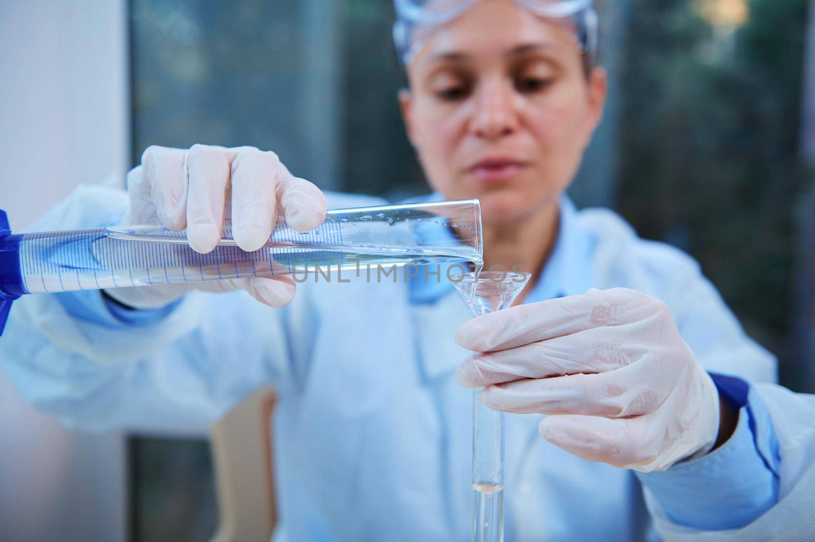 Details: Graduated cylinder in the gloved hands of a blurred woman scientist, lab technician pouring transparent liquid into a test tube through a laboratory funnel in a medical science laboratory