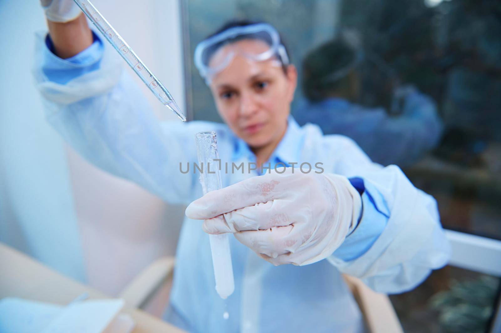 Selective focus on a test tube with a taking place chemical reaction and graduated lab pipette in a blurred woman scientist hands dripping reagent, conducting science experiment in research laboratory