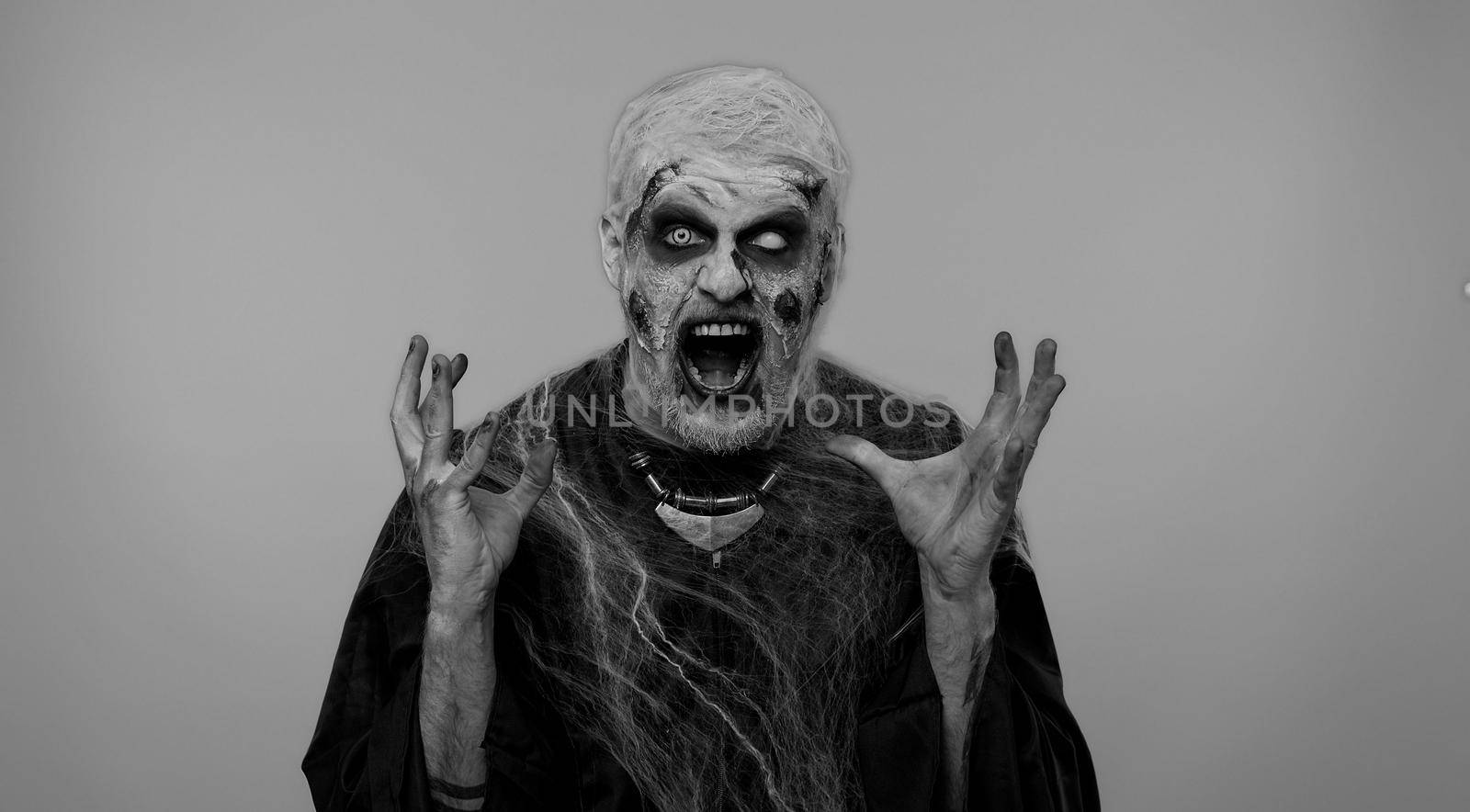 Scary man in carnival costume of Halloween crazy zombie with bloody wounded scars face screaming, shouting, trying to scare on gray. Horror theme of cosplay wounded undead, beast, monster
