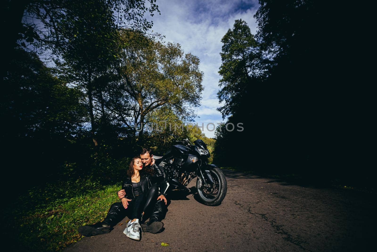 Handsome young man and beautiful young girl, couple sitting near motorcycle in black leather clothes, hugging, in nature, outdoors