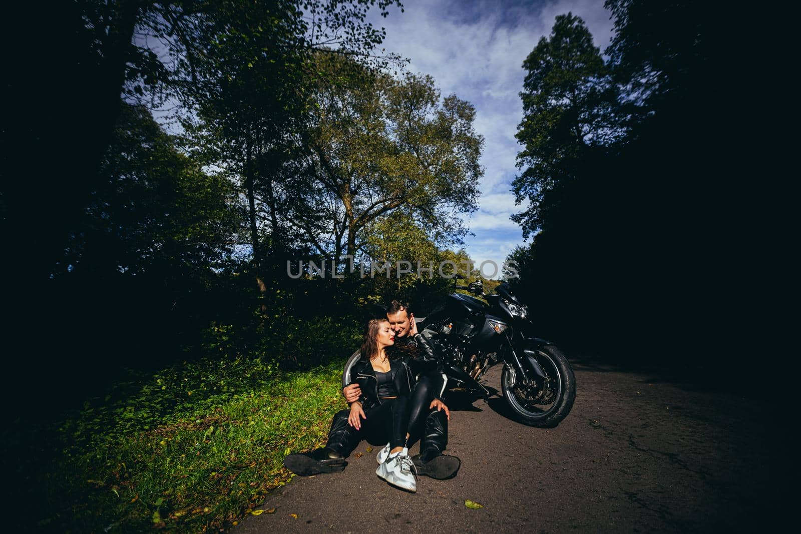 Handsome young man and beautiful young girl, couple sitting near motorcycle in black leather clothes, hugging, in nature, outdoors