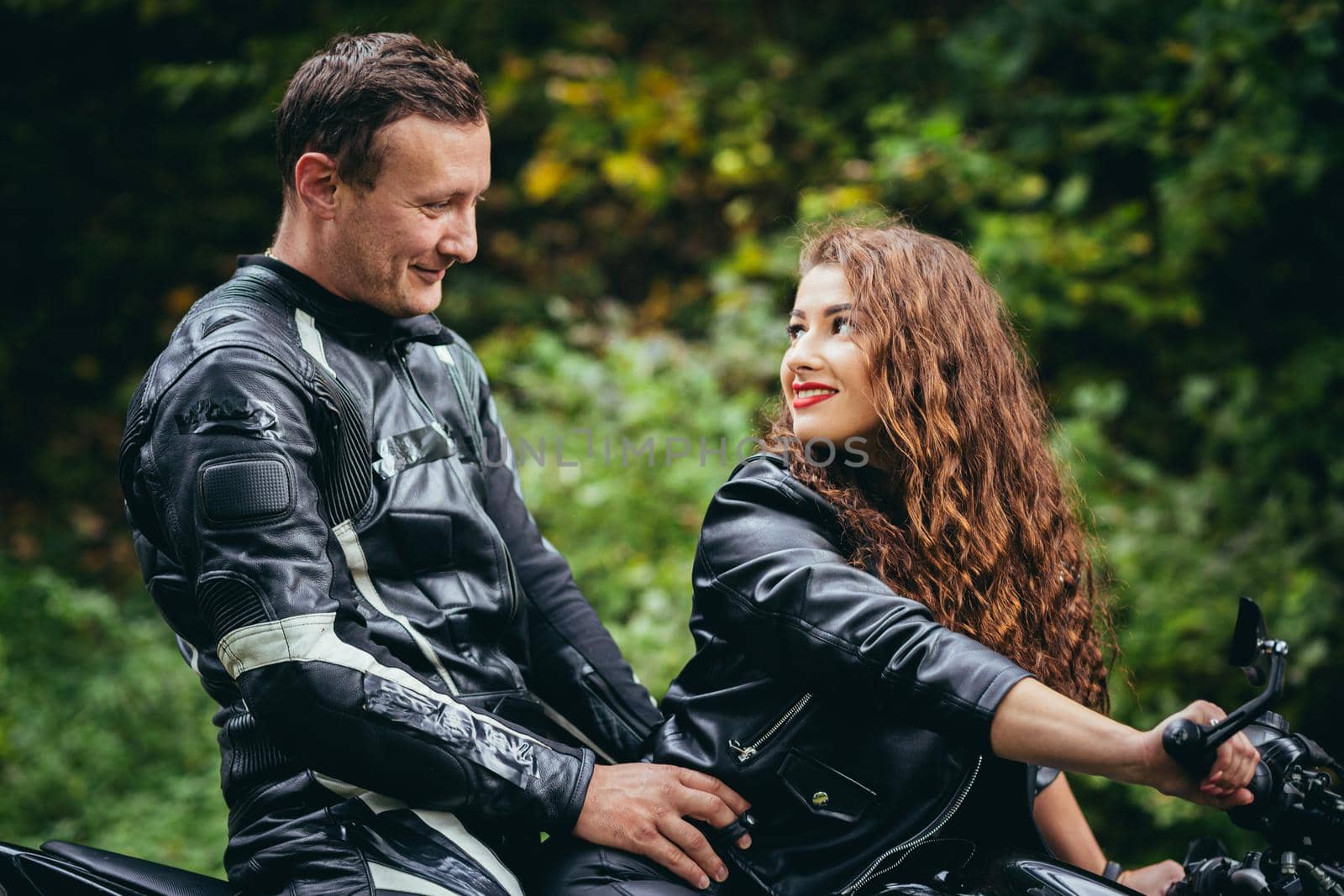 Handsome young man and beautiful young girl, couple sitting on a motorcycle in black leather clothes, close up, hugging, in nature, outdoors, looking at camera by voronaman