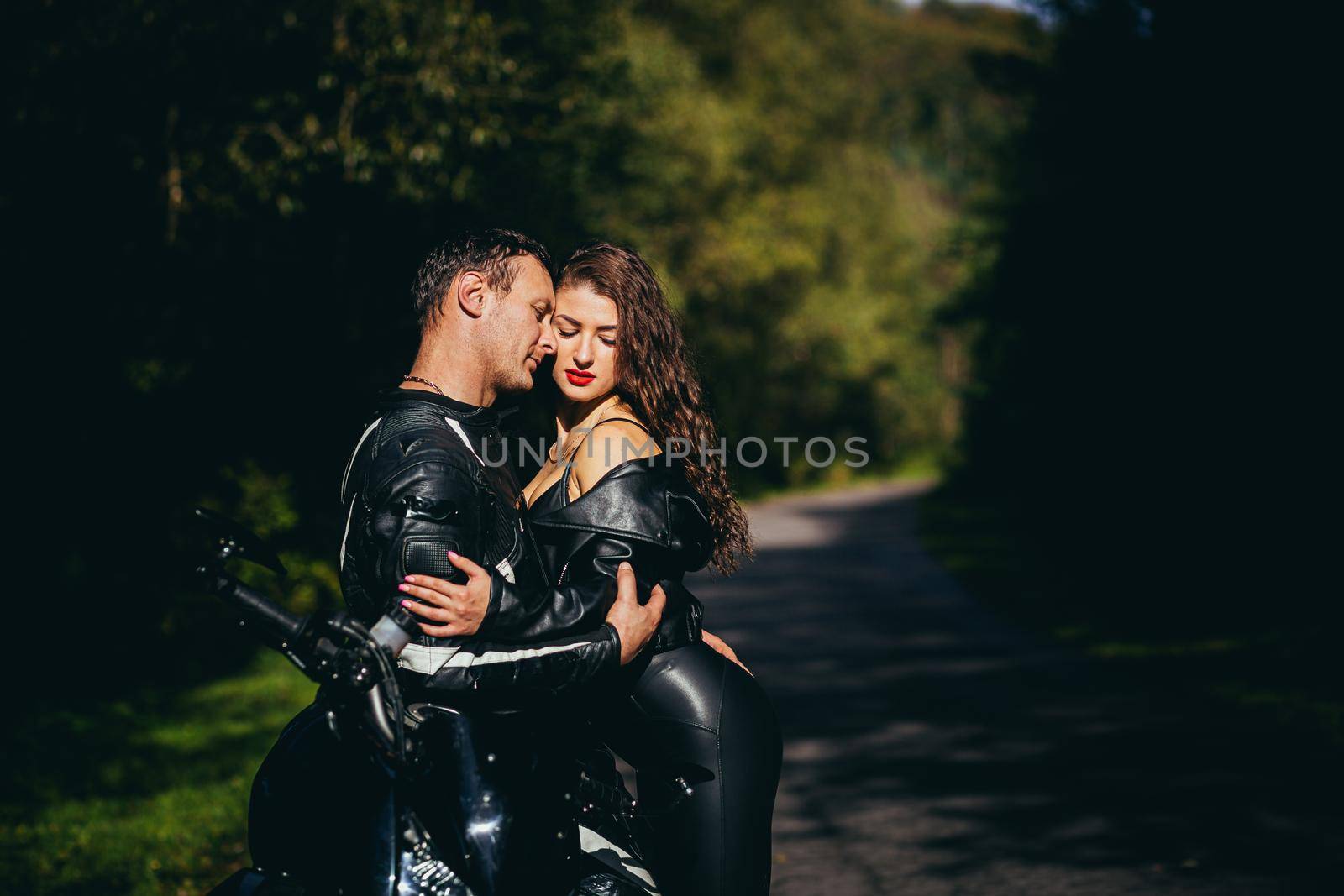 Handsome young man and beautiful young girl, couple sitting on a motorcycle, standing near a motorcycle in black leather clothes, hugging, in nature, on the street