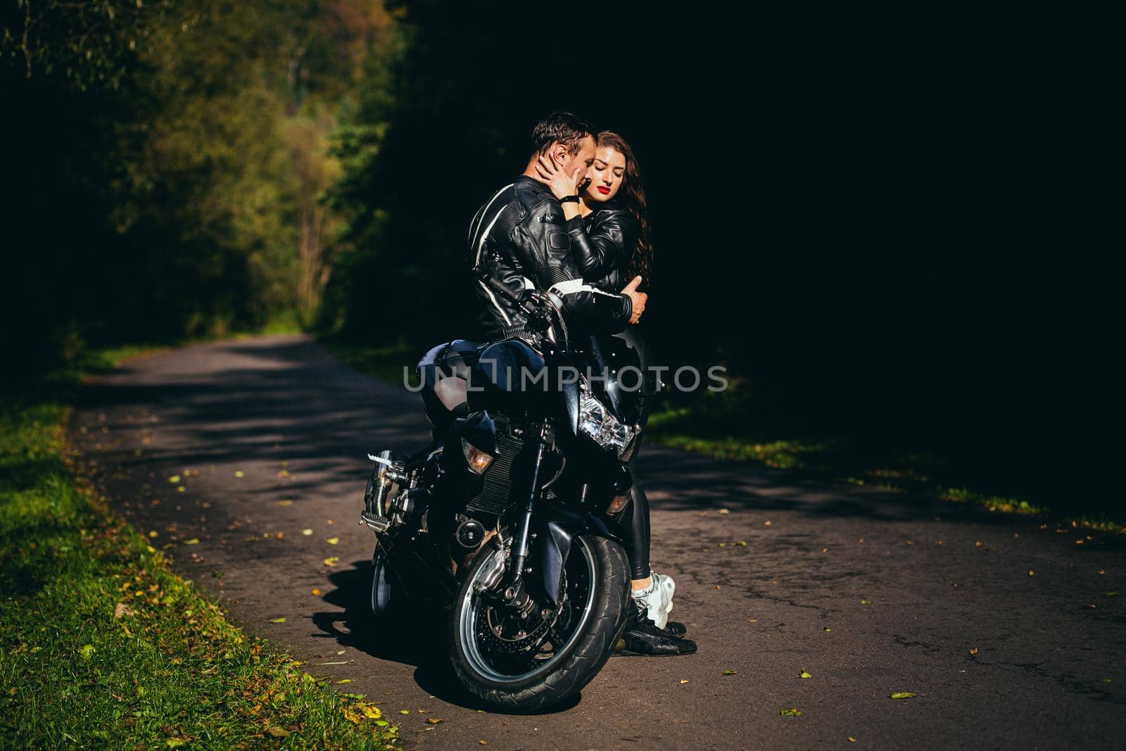 Handsome young man and beautiful young girl, couple sitting on a motorcycle, standing near a motorcycle in black leather clothes, hugging, in nature, on the street by voronaman