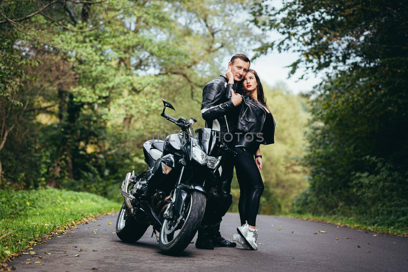 Handsome young man and beautiful young girl, couple sitting on a motorcycle, standing near a motorcycle in black leather clothes, hugging, in nature, on the street by voronaman