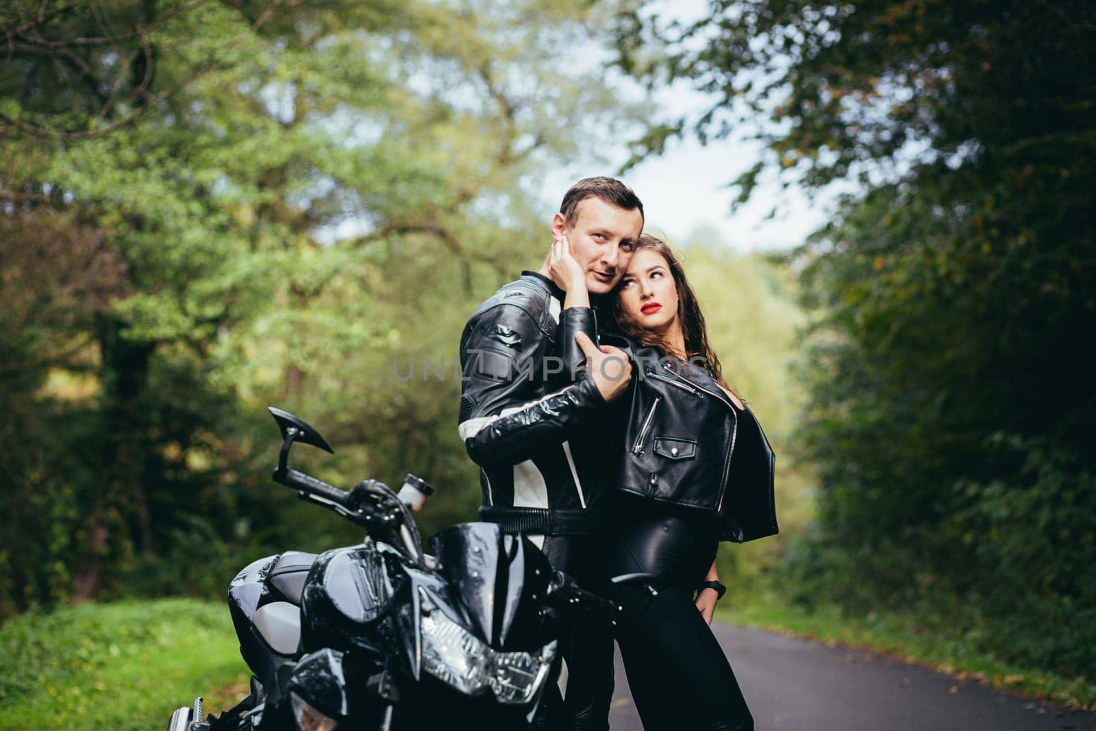 Handsome young man and beautiful young girl, couple sitting on a motorcycle, standing near a motorcycle in black leather clothes, hugging, in nature, on the street