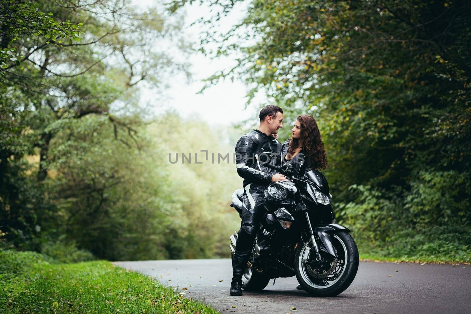 Handsome young man and beautiful young girl, couple sitting on a motorcycle, standing near a motorcycle in black leather clothes, hugging, in nature, on the street