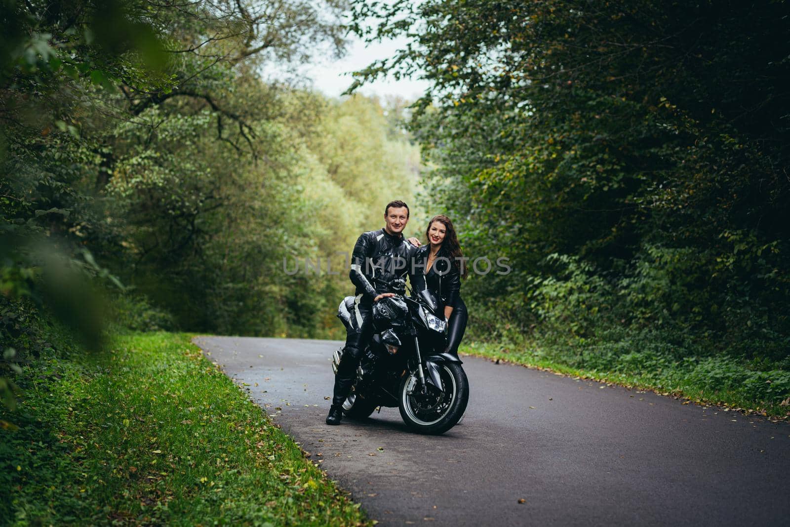 Handsome young man and beautiful young girl, couple sitting near motorcycle in black leather clothes, hugging, in nature, outdoors by voronaman